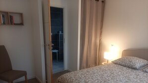 A bedroom with a bed, a chair, a bedside table with a lamp, a bookshelf, and a bathroom visible through an open door. - Jolie Maison Neuve, Très Bien Exposée, au Calme, Jardin, Terrasse (Sainte-Hélène)