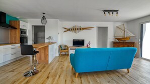A modern living room with a blue sofa, wooden flooring, a kitchen with a microwave and oven, a TV, and a sailboat decoration. - Jolie Maison Neuve, Très Bien Exposée, au Calme, Jardin, Terrasse (Sainte-Hélène)