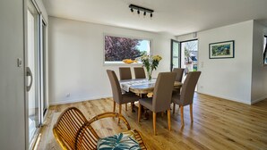 A dining area with a wooden table, six chairs, a vase with flowers, and a window with a view of trees and a yellow fish sculpture. - Jolie Maison Neuve, Très Bien Exposée, au Calme, Jardin, Terrasse (Sainte-Hélène)