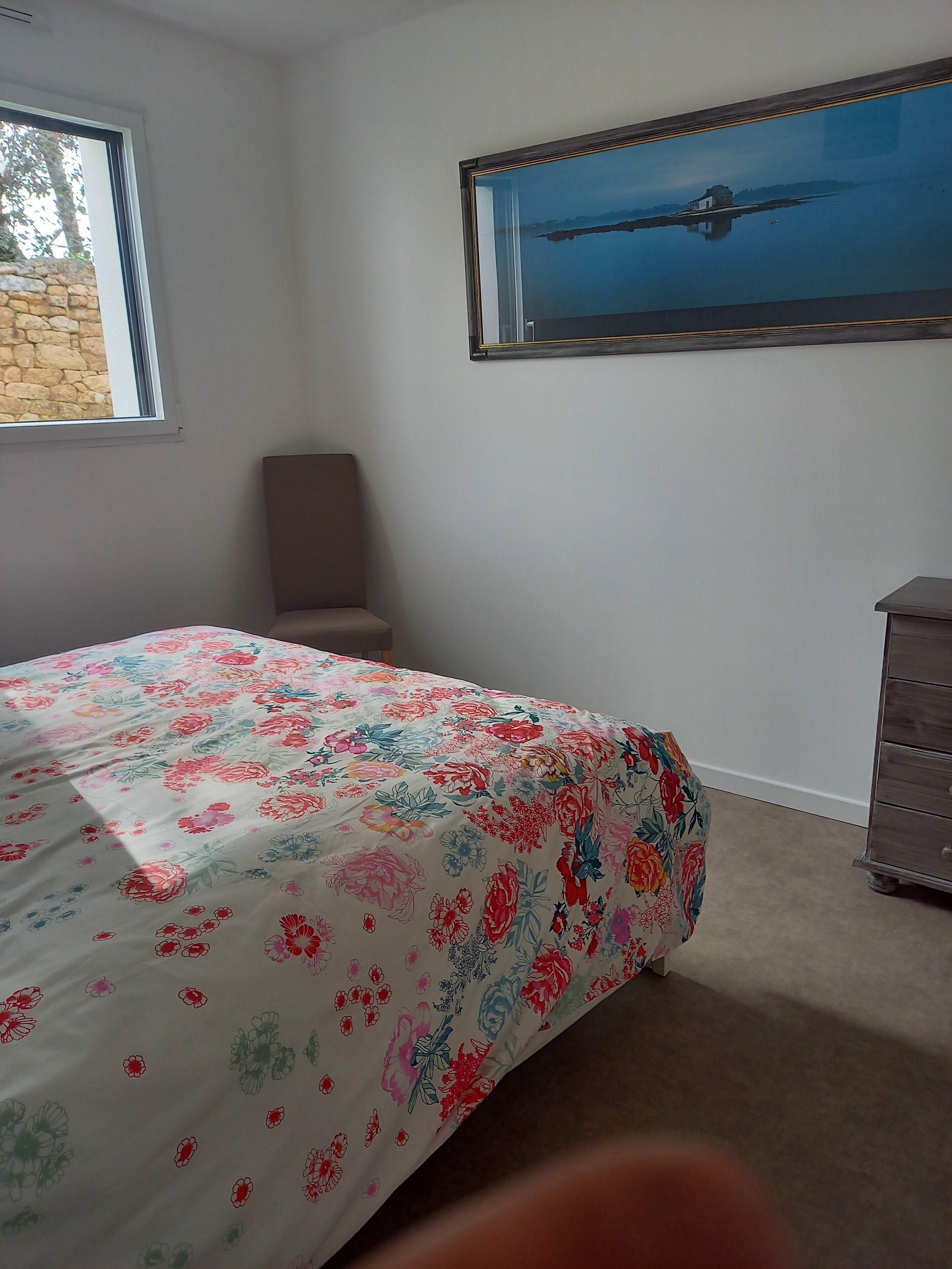 Un dormitorio con edredón estampado de flores, una ventana con vistas a un muro de piedra, una silla, un aparador y un cuadro en la pared.