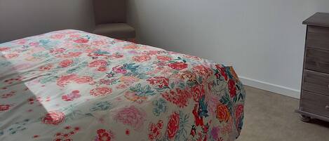 Un dormitorio con edredón estampado de flores, una ventana con vistas a un muro de piedra, una silla, un aparador y un cuadro en la pared.