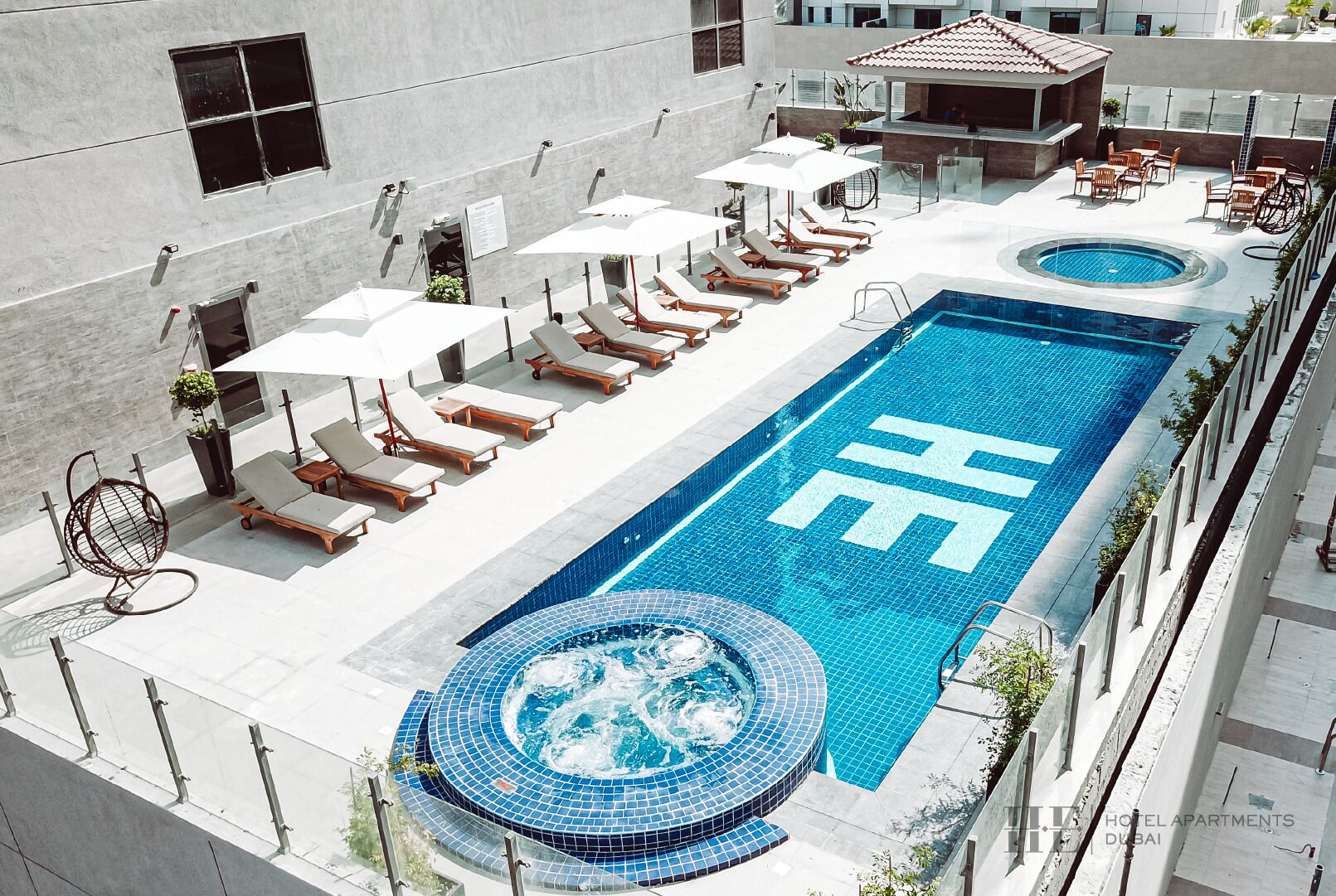 A rooftop pool area with lounge chairs and umbrellas, a hot tub, and a swimming pool with a large 'H' logo.