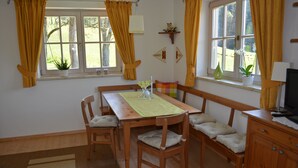 A dining area with a wooden table, chairs, and a bench. There are two windows with curtains, a small shelf with decorative items, and a television on a cabinet.