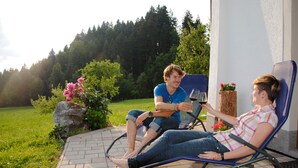 Two people sitting on outdoor chairs by a building with a garden and trees in the background.
