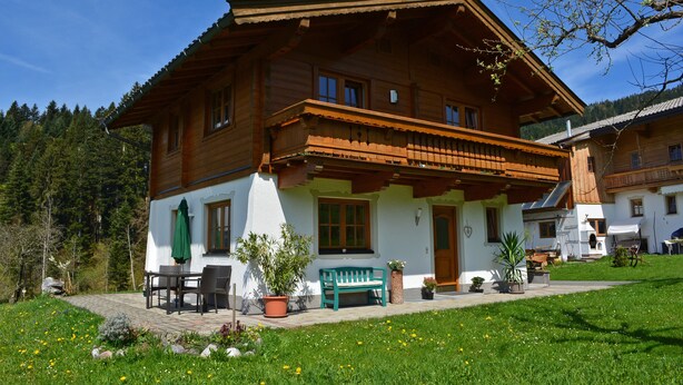 A wooden chalet with a balcony, a patio with a table and chairs, and a green bench.