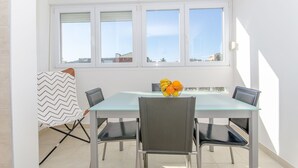 Een eethoek met een glazen tafel, vier stoelen, een fruitschaal en een raam met jaloezieën.