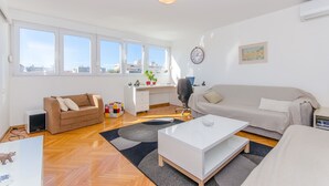 Een moderne woonkamer met een bank, een salontafel, een bureau en een computer. Grote ramen zorgen voor natuurlijk licht en uitzicht op gebouwen.