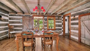 A rustic log cabin interior with a dining table set for four, wooden chairs, a chandelier, and a large window offering a view of greenery.
