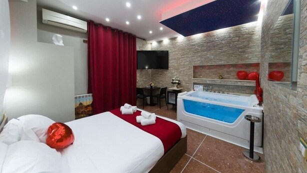 A hotel room with a large bed, a red curtain, a wall-mounted TV, a small table with chairs, a red decorative item, and a jacuzzi. - Cleopatra (Rome)