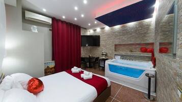 Camera d'albergo con un letto grande, una tenda rossa, una TV a parete, un tavolino con sedie, un elemento decorativo rosso e una vasca idromassaggio.