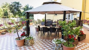 An outdoor patio with a checkered floor, a covered seating area with chairs and a table, and potted plants with flowers. - Cleopatra (Rome)