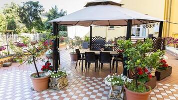 Una terrazza esterna con pavimento a scacchi, un'area salotto coperta con sedie e tavolo, e vasi con fiori.