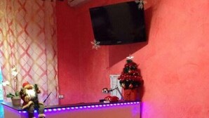A reception area with a red wall, a small Christmas tree, a TV mounted on the wall, and a reception desk with a purple light strip. - Cleopatra (Rome)