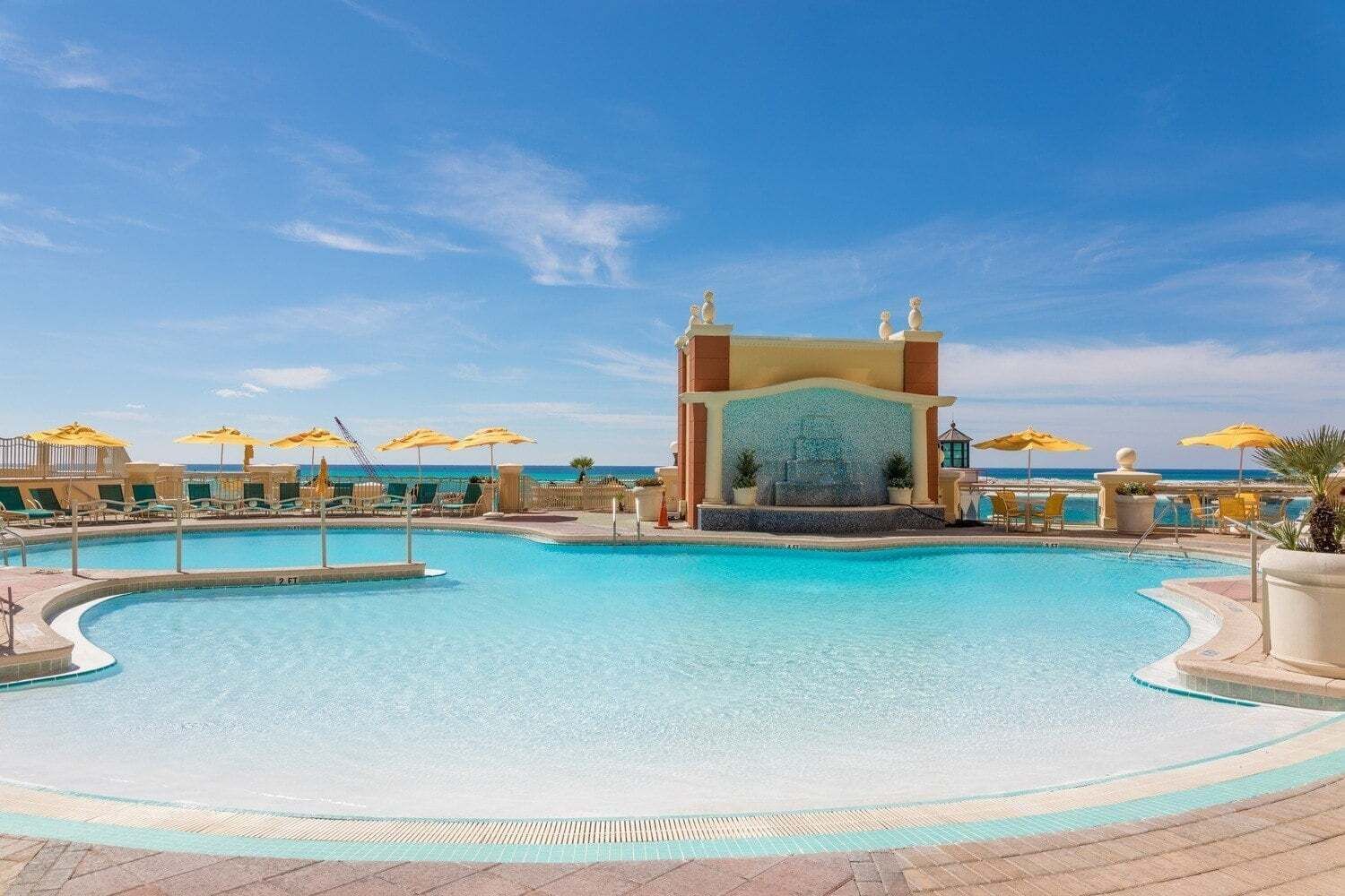 Une grande piscine extérieure dotée d’une fontaine centrale, entourée de chaises longues et de parasols.