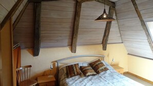 Un dormitorio con techo inclinado, vigas de madera, una cama con ropa de cama estampada, una mesita de noche, una silla y una lámpara colgante.