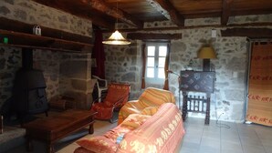 Una habitación rústica con paredes de piedra vista, techo de madera, un sofá con cojines estampados, una mesa de madera, una silla, una lámpara colgante y una chimenea.