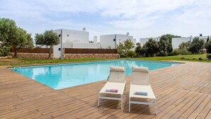 Une piscine avec deux chaises longues, une terrasse en bois et des bâtiments blancs en arrière-plan.