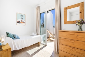 Un dormitorio con cama, cómoda de madera, espejo, cuadro enmarco y puerta corrediza de vidrio que da a un balcón.