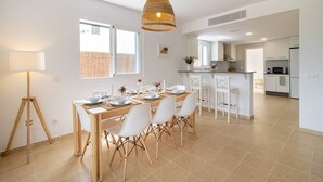 Une cuisine moderne avec des meubles blancs, un coin repas doté d’une table en bois et de chaises blanches, et une grande fenêtre qui laisse entrer la lumière naturelle.
