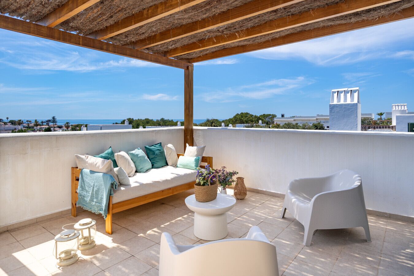 A rooftop terrace with a wooden pergola, a white sofa with cushions, a white armchair, and a small table with a vase of flowers.