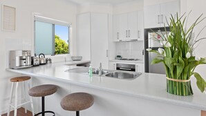 Een moderne keuken met witte kasten, een roestvrijstalen spoelbak en een ingebouwde koffiezetapparaat. Op het aanrecht staan twee krukken en een vaas met groene planten.