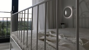 A bedroom with a white bed, a round mirror, and a view of greenery outside.