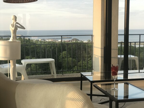 Varanda com sofá branco, uma mesa pequena e uma estátua. A vista inclui um corpo d'água e vegetação.