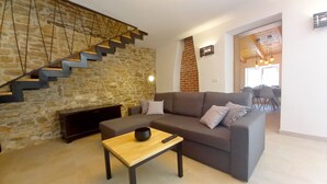 Una sala de estar con una pared de piedra, un sofá gris, una mesa de centro de madera y una escalera con barandilla de madera.