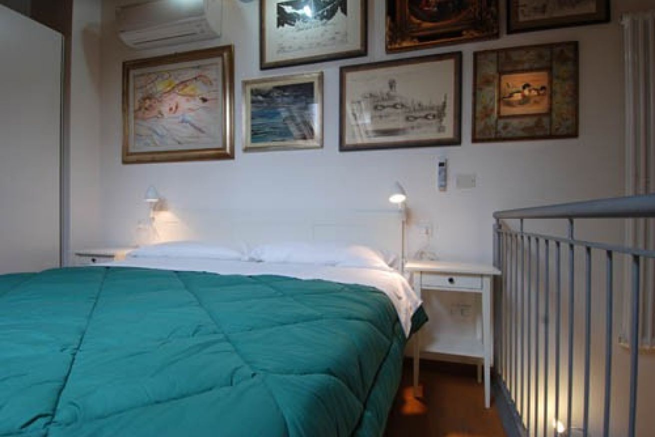 Een slaapkamer met een groene sprei, een nachtkastje met een lamp en een muur versierd met ingekaderde foto's.