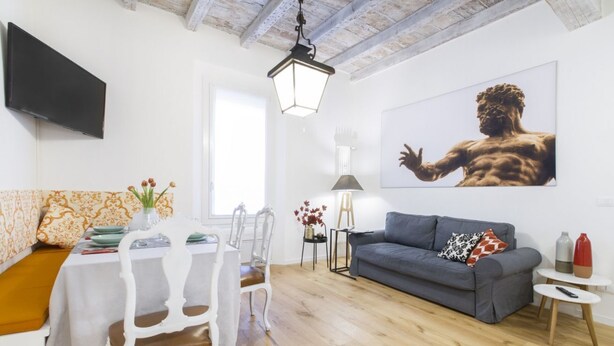 A modern living room with a dining area, a grey sofa, a TV mounted on the wall, and a large painting of a muscular figure. - GetTheKey San Vitale Apartment (Bologna)