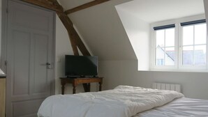 A bedroom with a sloped ceiling, a bed with white bedding, a television on a small wooden table, and a window with a view of buildings. - Renovated house 100m from sea with WIFI, garden in Trégastel (Trégastel)
