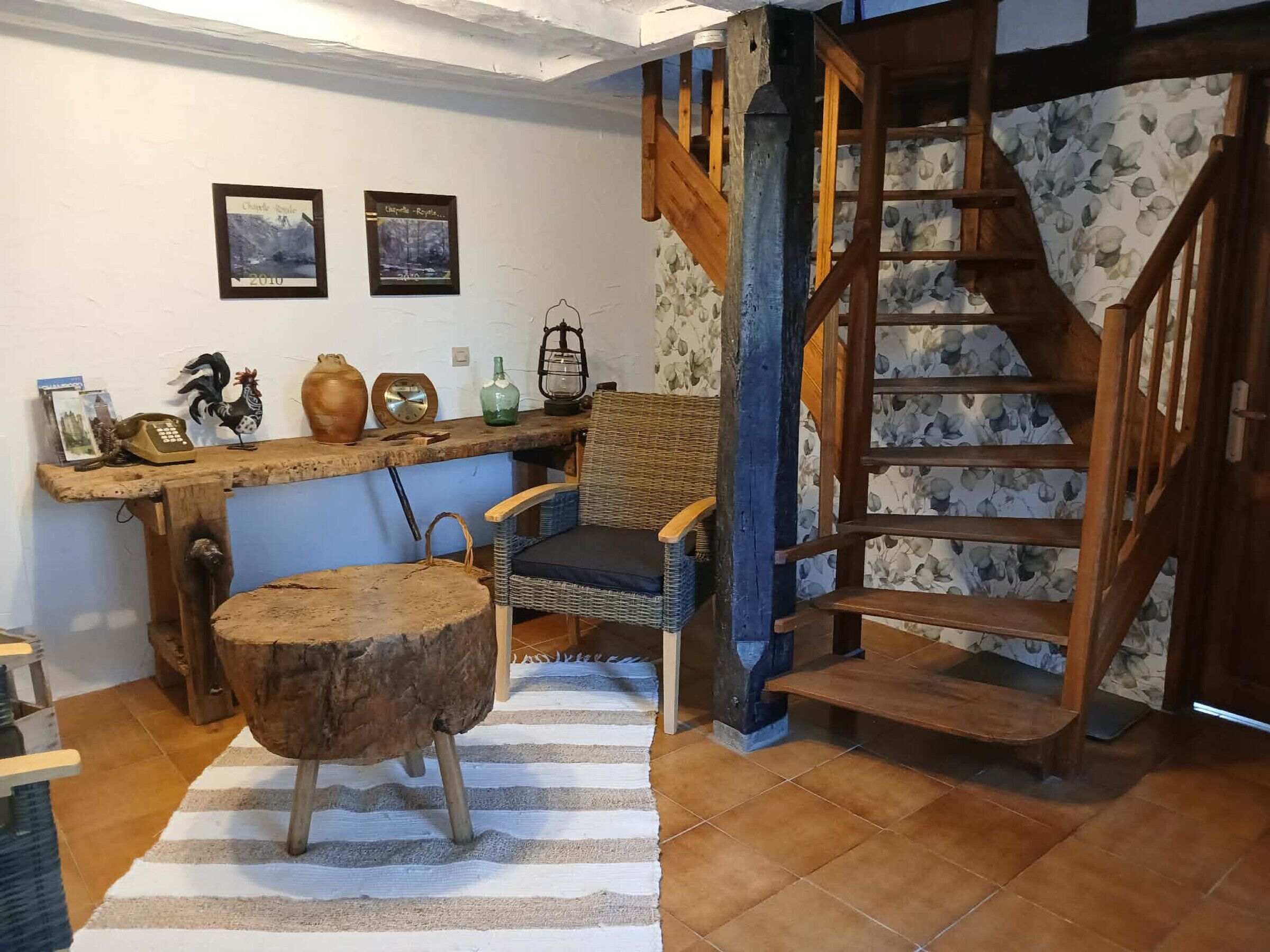 A rustic interior with a wooden staircase, a wicker chair, a wooden stool, and a wall-mounted shelf with various items.