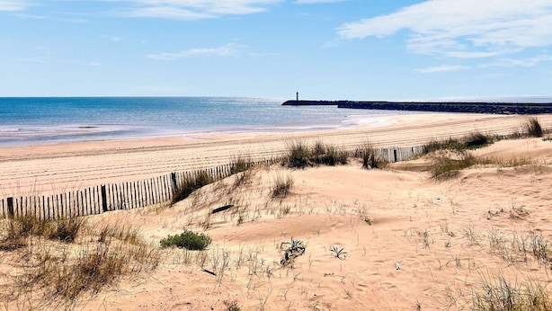 Una playa con una valla de madera, dunas arenosas y un faro a lo lejos.