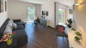 Ein modernes Wohnzimmer mit einer Couch, einem kleinen Tisch und einem Fernseher. Ein Balkon mit Blick auf Bäume und einem Geländer ist vorhanden.