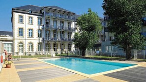 A swimming pool with a wooden deck, surrounded by a building with multiple balconies and a large tree.
