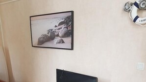 A living room with a flat-screen TV on a white entertainment unit, a small side table with magazines, and a framed picture of a rocky beach on the wall.