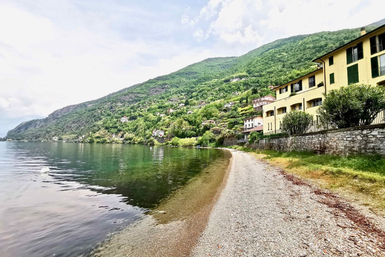 A coastal road with buildings on a hillside, a clear lake, and a stone wall.