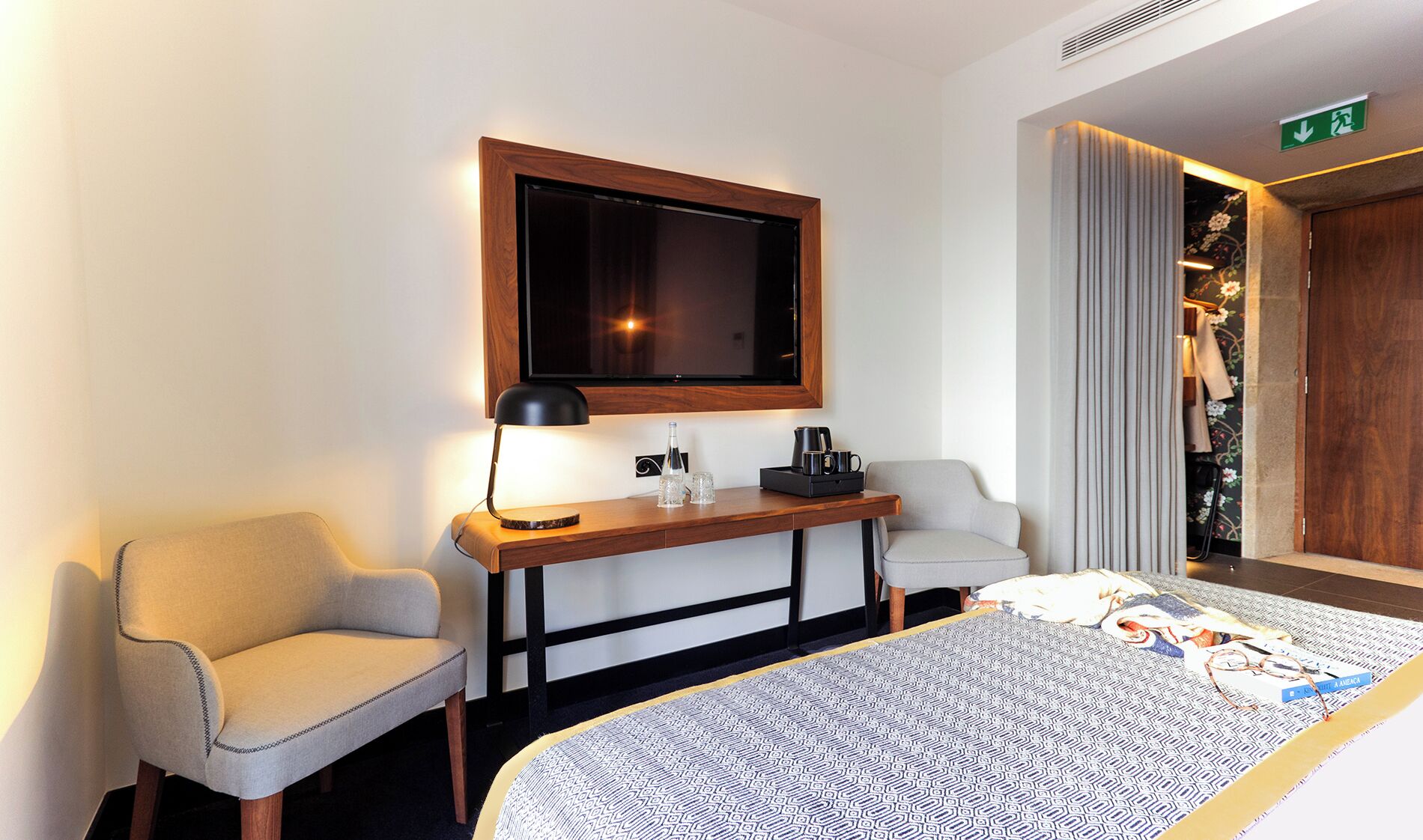 A hotel room with a bed, two armchairs, a desk with a lamp, a television, and a door with a floral wallpaper.
