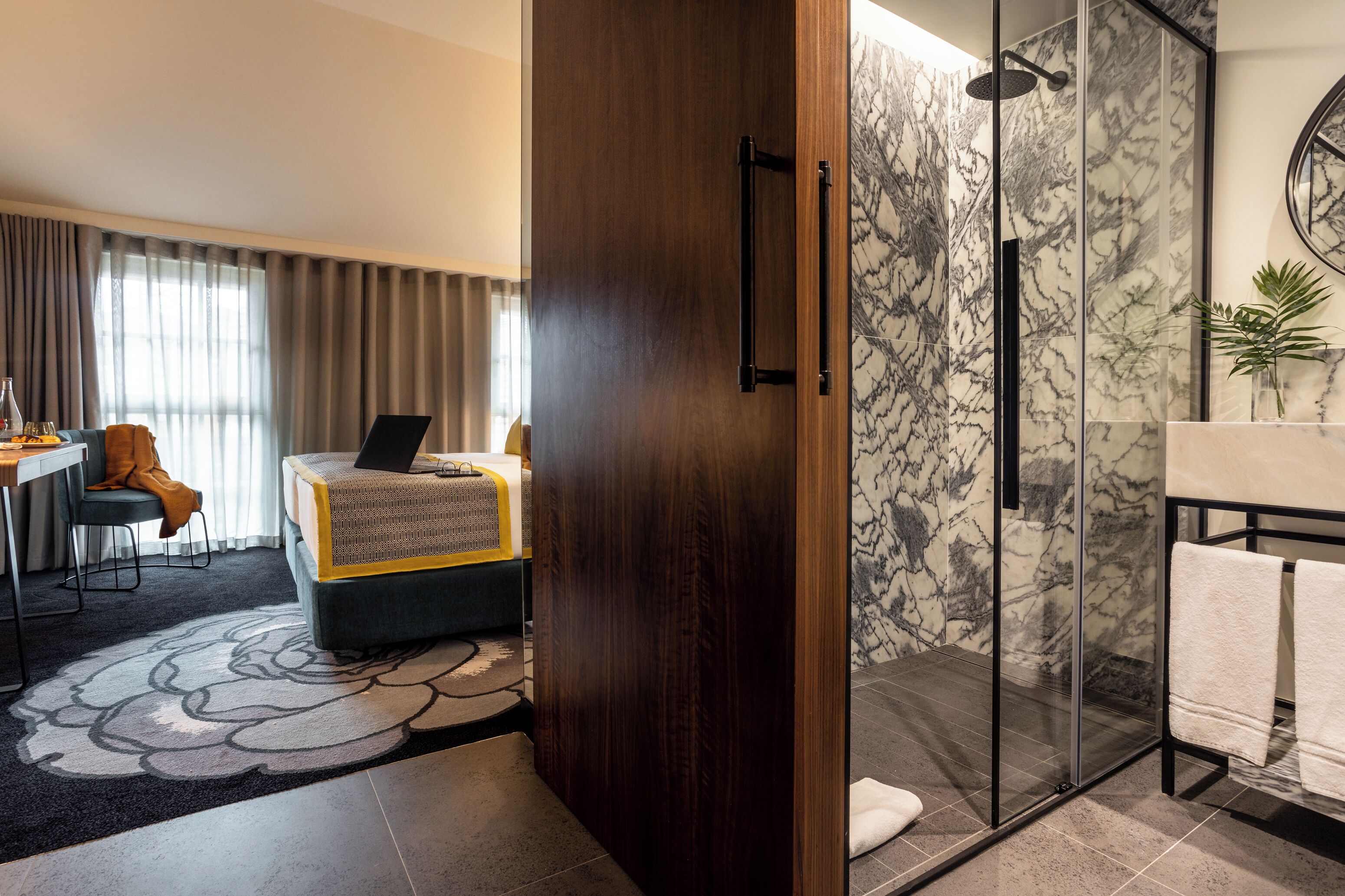 A modern hotel room with a wooden door, a glass shower enclosure, a marble-tiled shower area, a small seating area with a laptop, and a dining table with a chair.