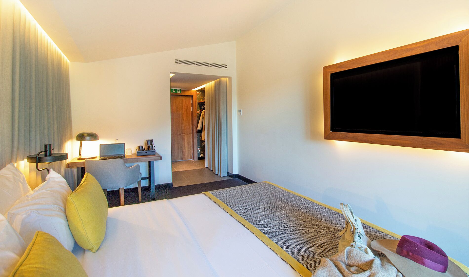 A hotel room with a bed, a desk with a laptop, a chair, and a television mounted on the wall.