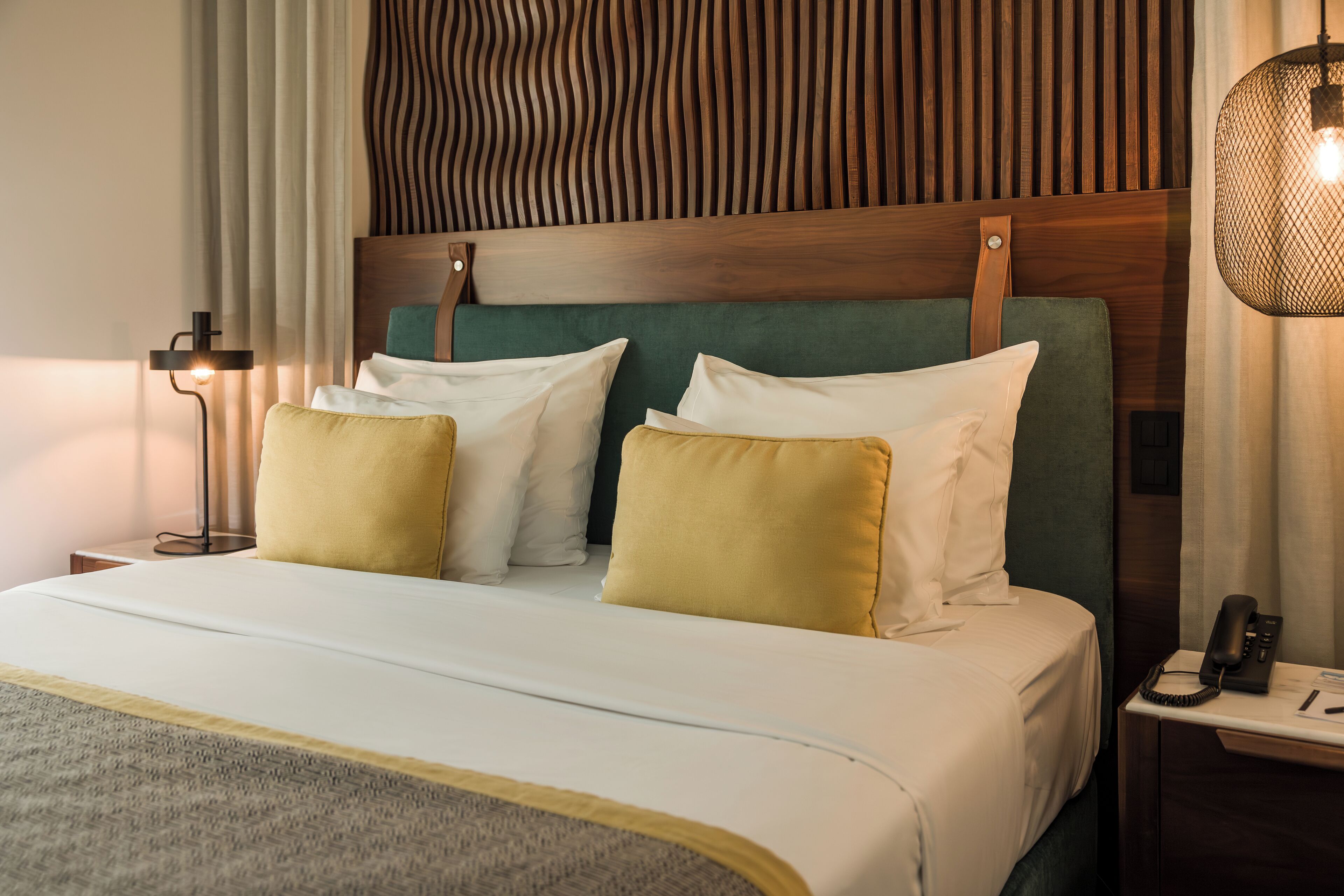 A neatly made bed with white and yellow pillows, a green headboard, and a bedside table with a lamp and a phone.