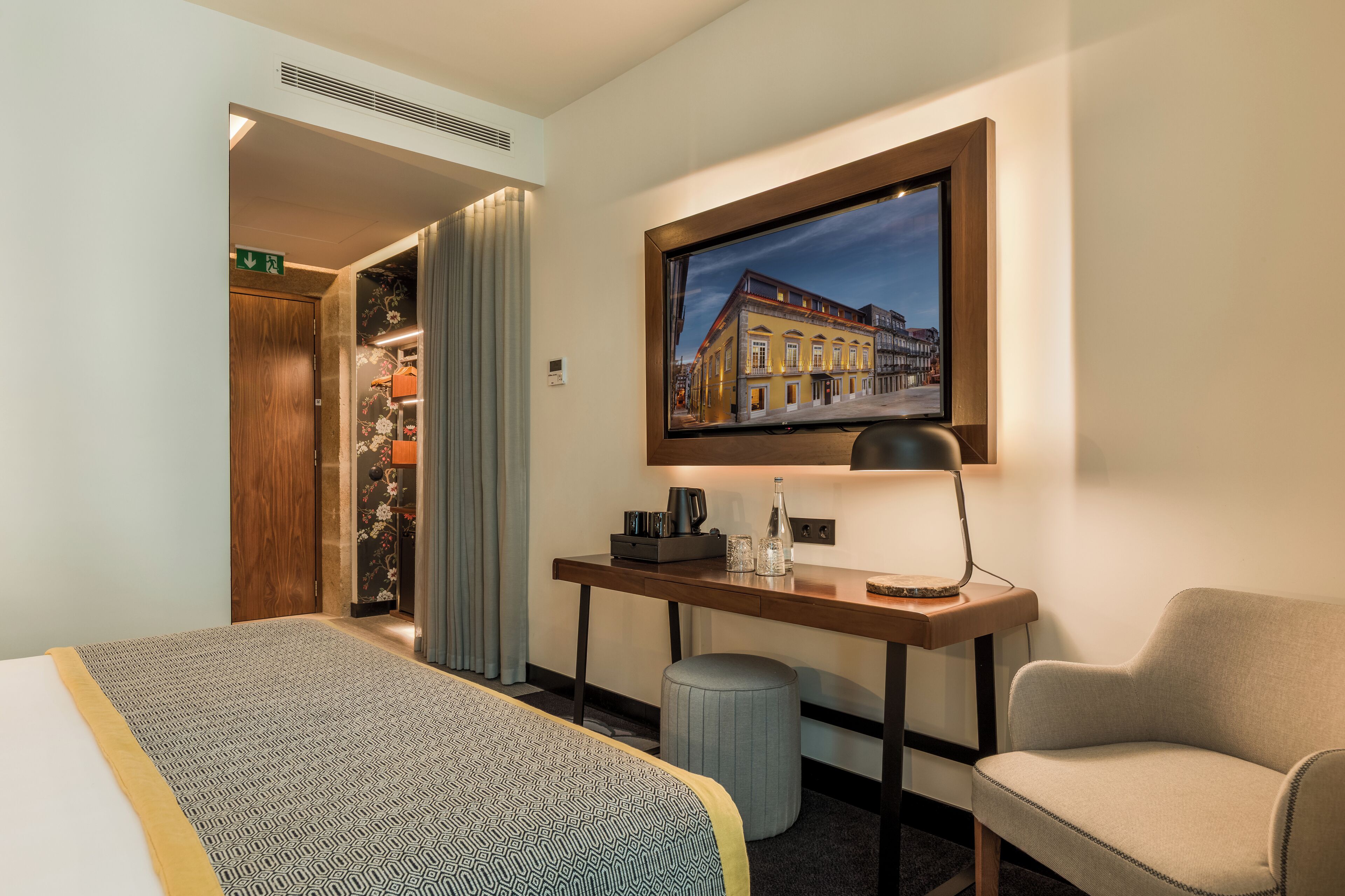 Habitación de hotel con cama, escritorio con lámpara, una silla, televisor montado en la pared y un cuadro con una imagen de un edificio.