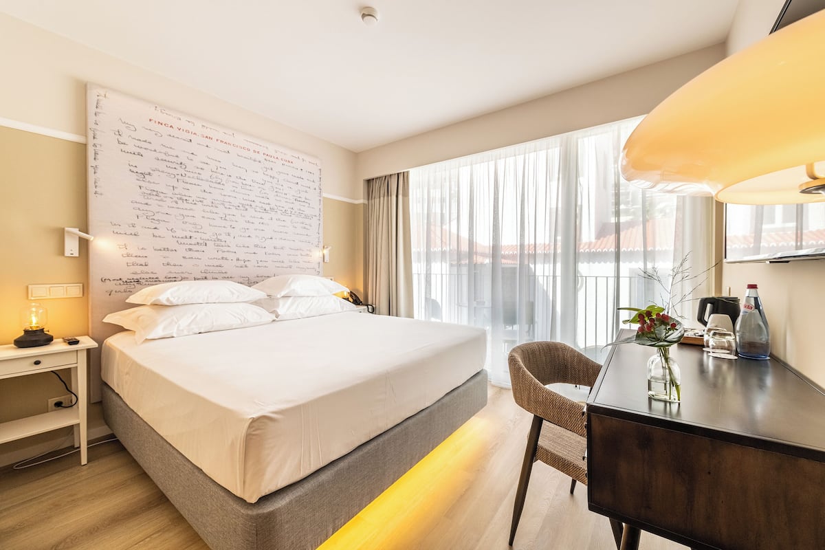 Ein Hotelzimmer mit einem großen Bett, einem Schreibtisch mit Lampe, einem Sessel und Blick auf die Stadt durch große Fenster.