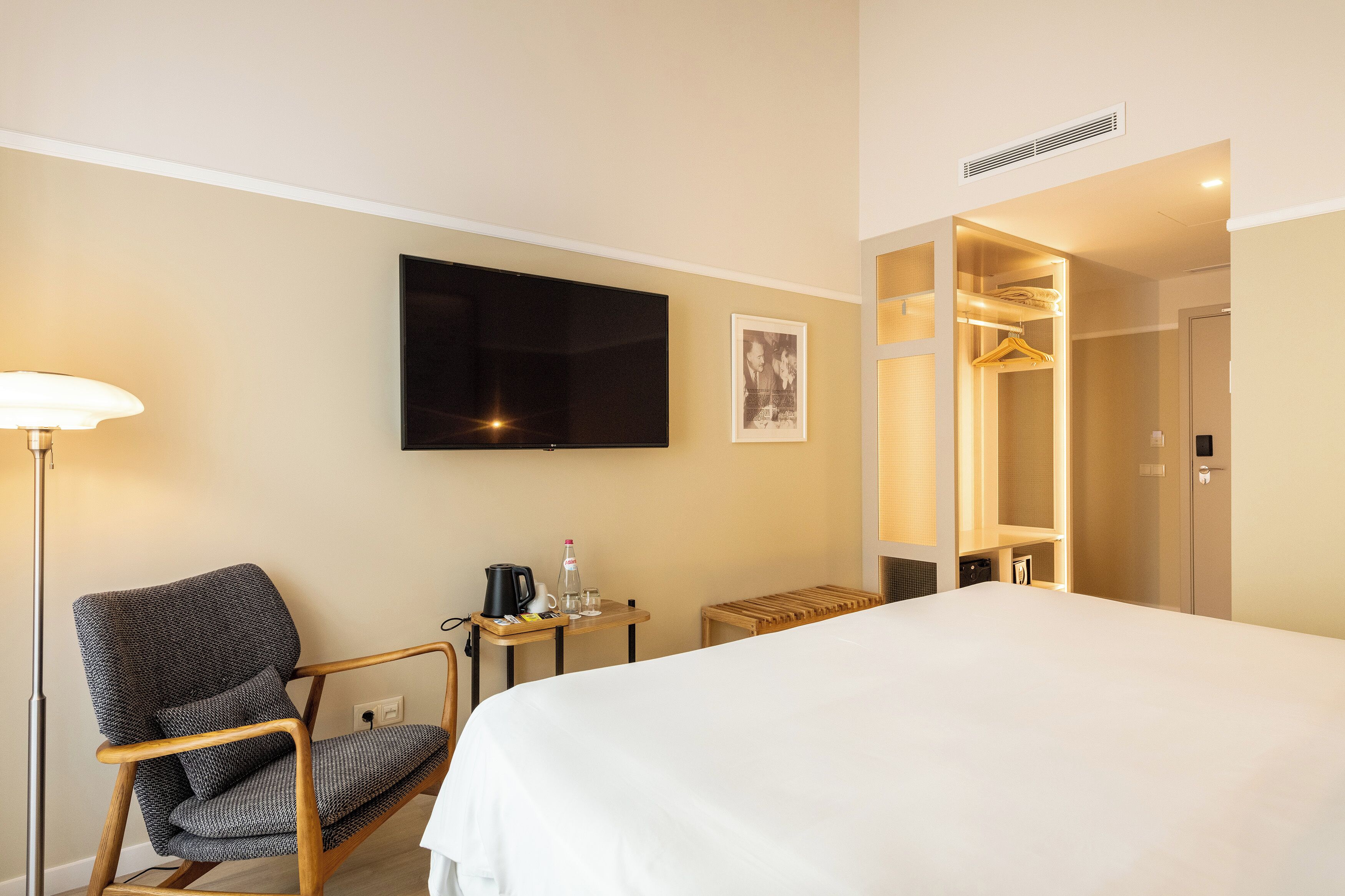 A hotel room with a bed, a chair, a TV, a bedside table with a kettle and glasses, and a wardrobe.