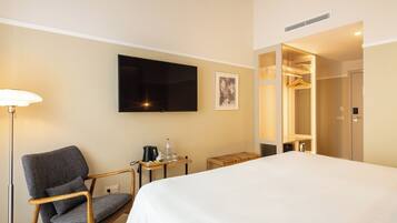 A hotel room with a bed, a chair, a TV, a bedside table with a kettle and glasses, and a wardrobe.