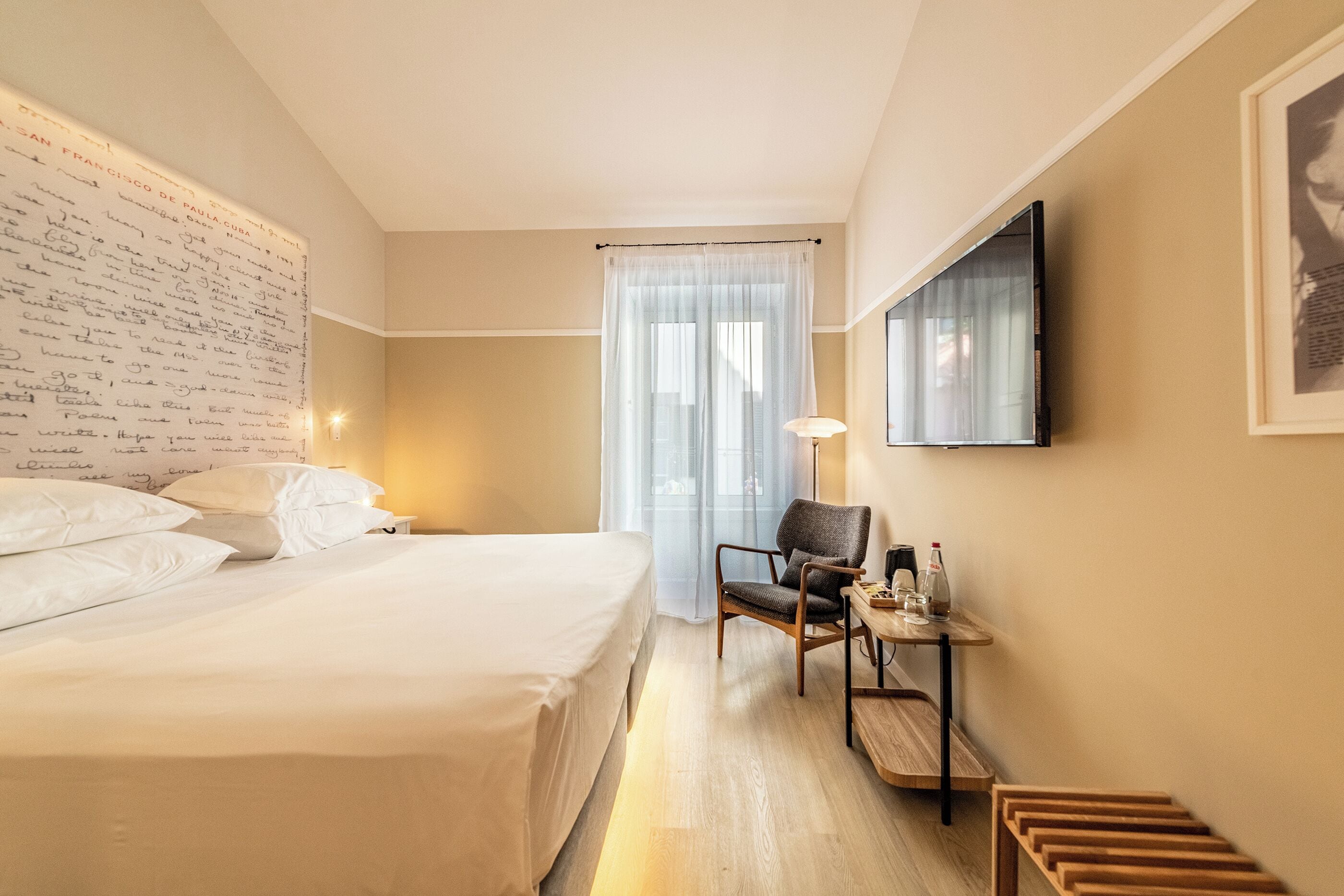 Habitación de hotel con una cama grande, un escritorio con silla, televisión y ventana con cortinas translúcidas.