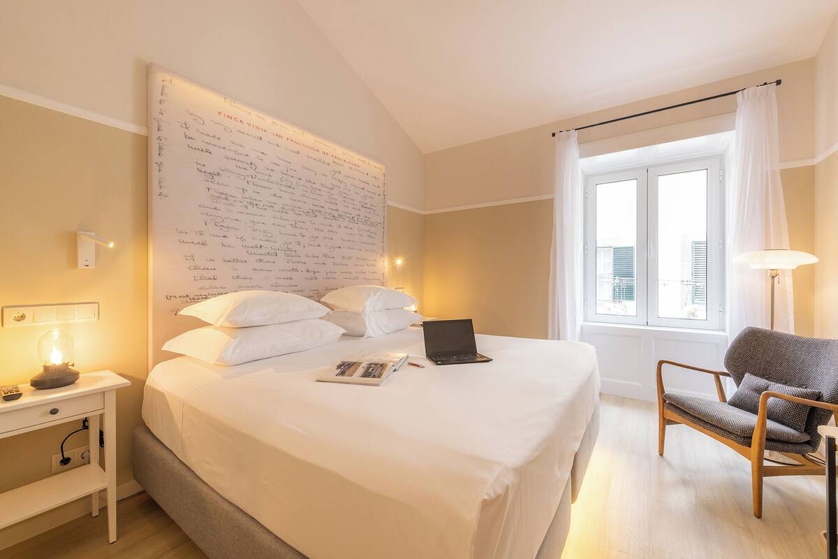 Ein Hotelzimmer mit einem großen Bett, einem Laptop, einem Sessel und einem Fenster mit Vorhängen.