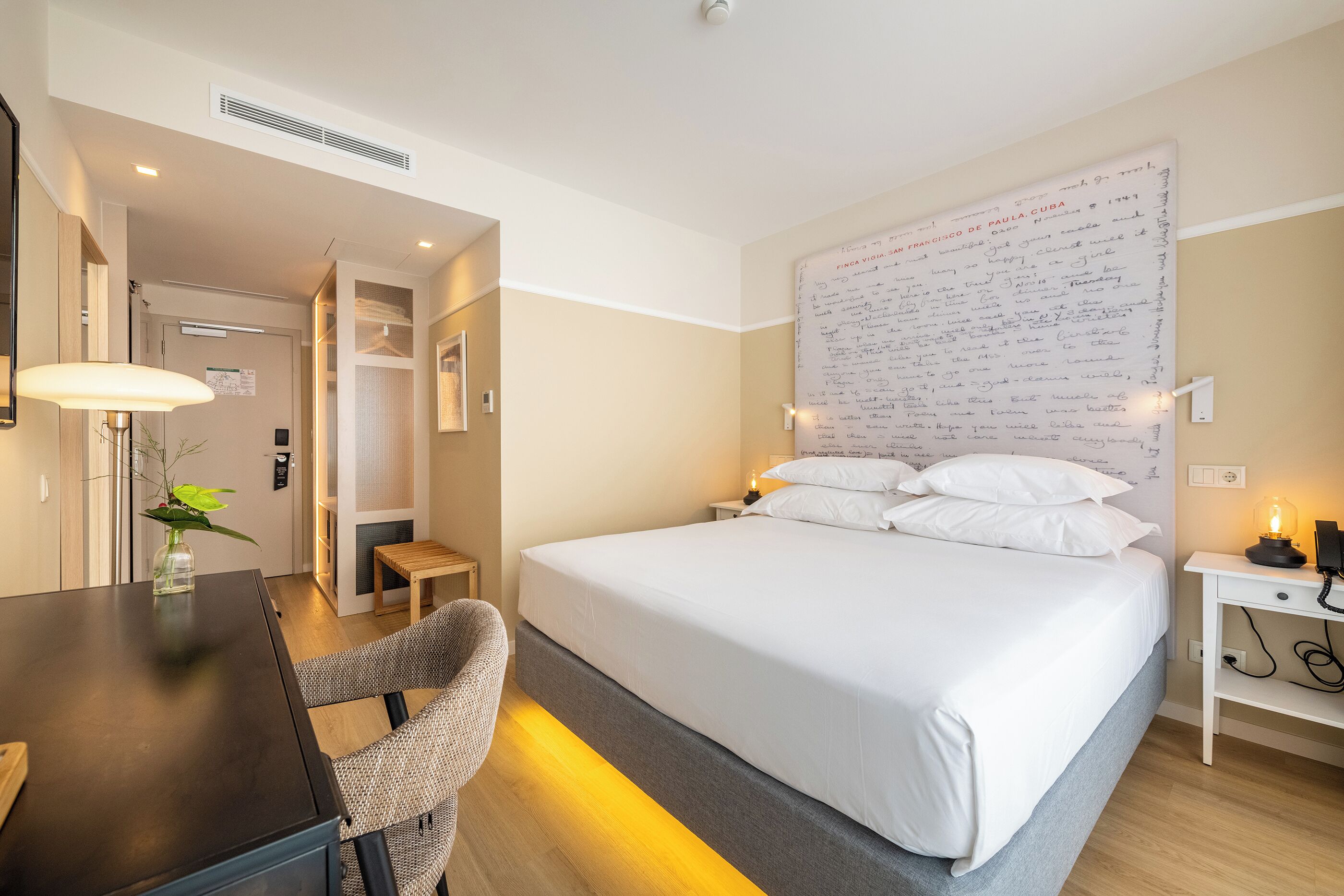 Una habitación de hotel con cama, mesita de noche, lámpara, silla y una pared con notas escritas a mano.