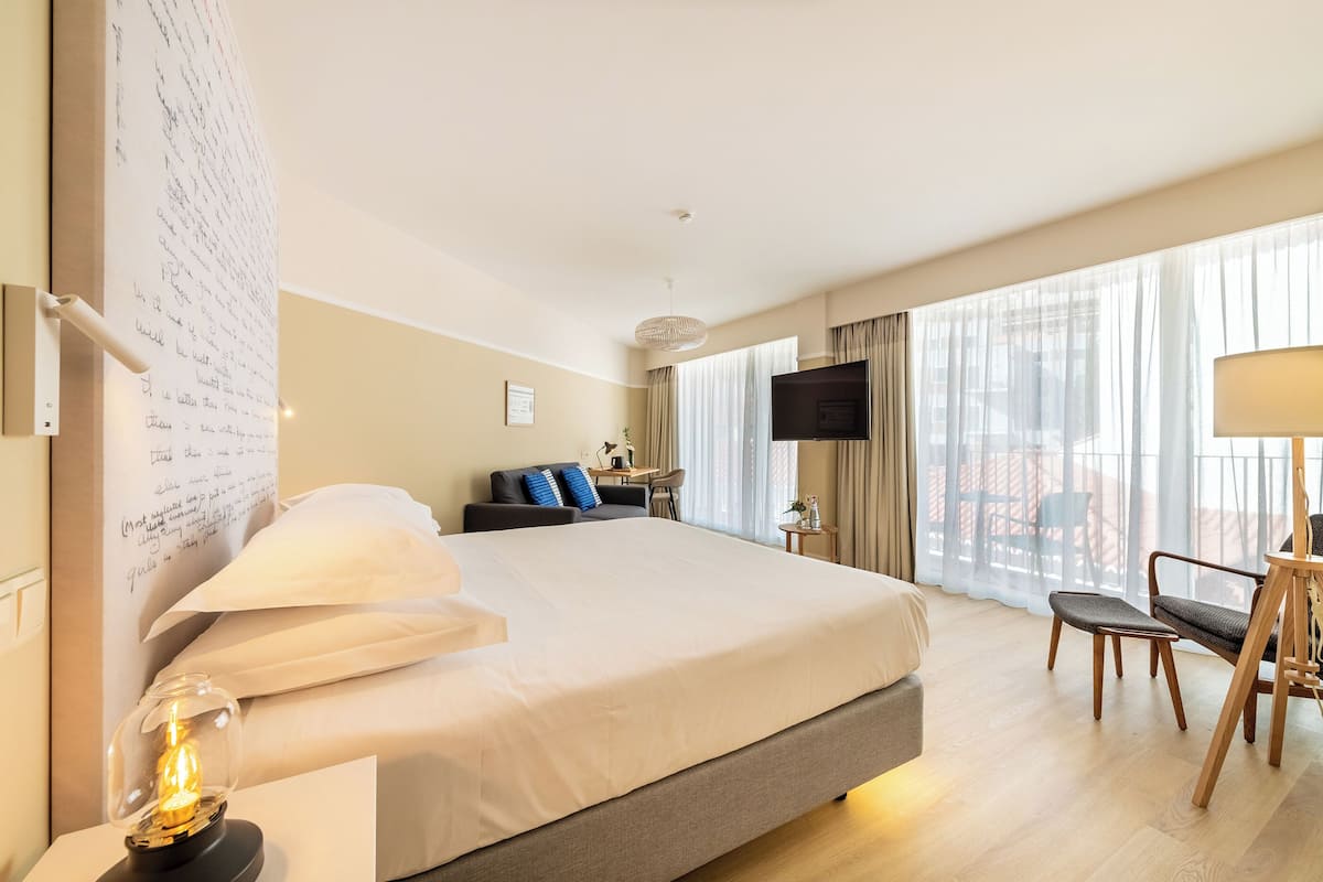Ein Hotelzimmer mit einem großen Bett, einem Schreibtisch mit Stuhl, einem Fernseher und einem großen Fenster mit Vorhängen.