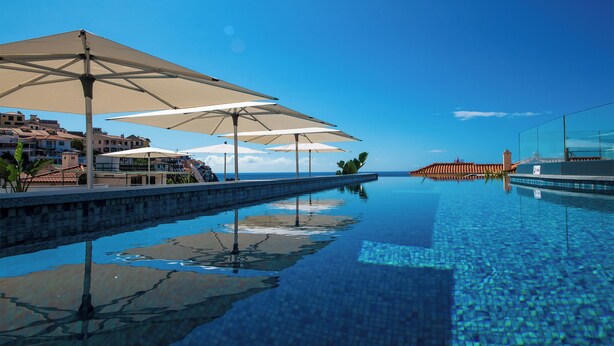 A swimming pool with clear blue water, surrounded by lounge chairs and umbrellas, and buildings in the background. - Pestana Fisherman Village (Camara de Lobos)
